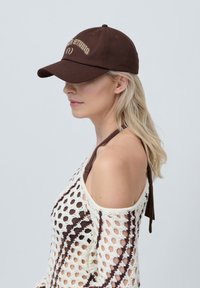 Brown baseball cap with embroidered "Studio" in beige, featuring a curved brim and adjustable strap at the back.