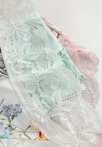 Fine lingeristykker med hvidt blomstret blonder, pastelgrønne og lyserøde stoffer samt et tøjstykke med blomsterprint under.