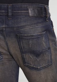 Jeans de mezclilla azul oscuro desgastados con detalles rasgados, diseño de cinco bolsillos y parche de cuero con logo en la cintura trasera.