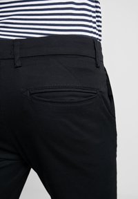 Pantaloni neri con una texture liscia e una tasca posteriore con un'apertura curva, abbinati a una camicia a righe blu e bianche in vita.