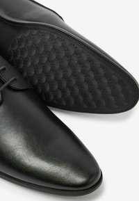 Zapato de vestir de cuero negro con punta afilada y suela de goma texturizada con un patrón scallopado. Incluye cordones, diseño minimalista.