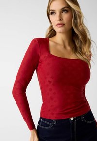 Rode top met lange mouwen en een vierkante halslijn, gemaakt van zachte stof met een subtiel tonale reliëfpatroon. Gecombineerd met donkere spijkerbroek.