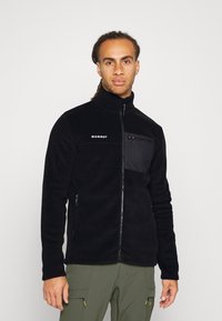 Black fleece jacket with high collar, zipper front, and chest pocket. Features Mammut logo and side zippered pockets.
