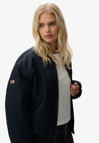 Marine bomberjack met een gladde, glanzende textuur. Heeft een zichtbare ritssluiting en een logopatch op de linker mouw. Draagt over een witte top.
