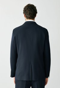 Uomo con capelli scuri e ricci, indossa un abito blu navy e una camicia bianca, in piedi di spalle contro uno sfondo bianco uniforme.