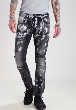 Mand iført sorte slidte jeans med hvide malingstænk, revner ved knæene, sorte sneakers og en lysegrå skjorte, stående mod en hvid baggrund.