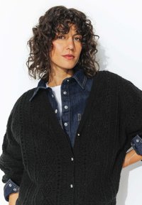 Cardigan noir en tricot avec un motif texturé, doté de grands boutons. Porté sur une chemise en denim foncé avec un col et des poignets contrastants.