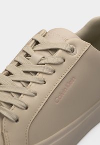 Μπεζ αθλητικό παπούτσι Calvin Klein με υφασμένους κορδόνια, ανάγλυφο λογότυπο κοντά στη φτέρνα και λείο συνθετικό επάνω μέρος σε κοντινό πλάνο.
