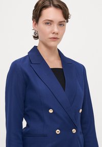 Kaksirivinen tummansininen bleiseri, jossa on harpoinikuvio, kultaiset napit, terävät kaulukset ja kaksi etutaskua. Yksinkertainen, rakenteellinen muotoilu.