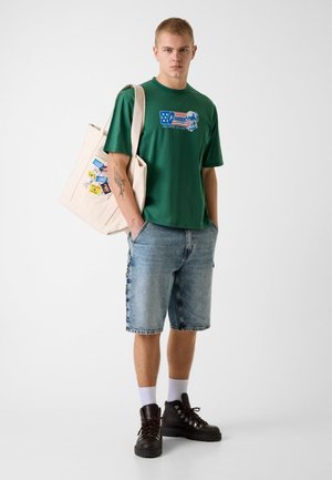 Młody mężczyzna w zielonym t-shircie z nadrukiem, dżinsowych szortach, czarnych butach, białych skarpetkach, niosący beżową torbę na ramieniu z kolorowymi naszywkami.