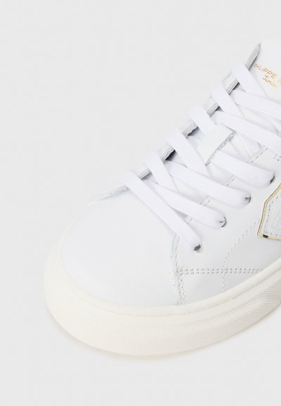 Weiße Ledersneaker mit flachen weißen Schnürsenkeln, strukturieter weißer Gummisohle und einem grauen Akzent an der Seite. Verfügt über ein goldfarbenes Logodesign.