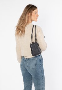 Piccola borsa nera in pelle con una texture liscia, forma rettangolare, tracolla corta regolabile e design minimalista. Indossata sopra un maglione beige.