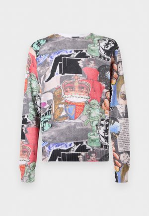 Jonge man draagt een kleurrijke trui met een collage van kronen, beelden en gezichten, gecombineerd met lichtblauwe jeans tegen een effen achtergrond.