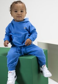 Blauwe katoenen hoodie met een voorzak en bijpassende joggingbroek; witte Nike sneakers met zwarte swoosh. Kind zit op groene blokken.