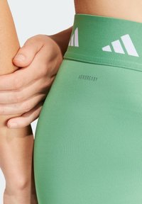 Gröna leggings med en slät textur och hög midja. Har vita logotyper och texten "AEROREADY", utformad för att hantera fukt.