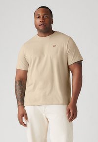Beige, rövid ujjú póló puha pamutból, kerek nyakkivágással és egy kis piros logóval a mellkasán, fehér nadrággal párosítva.