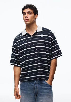 Mladý muž v čierno-bielom pruhovanom polotričku a modrých džínsoch, mierne sa pozerá do strany s jednou rukou v vrecku.