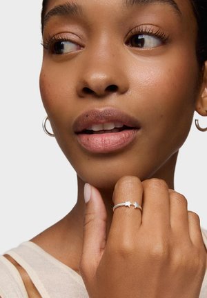 Zbliżenie na kobietę dotykającą podbródka, noszącą srebrny pierścionek z dwoma gwiazdkami oraz małe kolczyki koła.