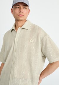Beige short-sleeve shirt featuring a perforated design, button front, and a small logo on the left chest. Light, airy fabric texture.