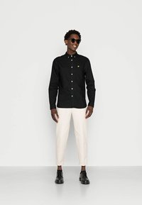 Μαύρο πουκάμισο με κουμπιά και ένα μικρό κίτρινο λογότυπο, συνδυασμένο με κρεμ παντελόνια σε tapered γραμμή και μαύρα παπούτσια, φορεμένο με γυαλιά ηλίου.