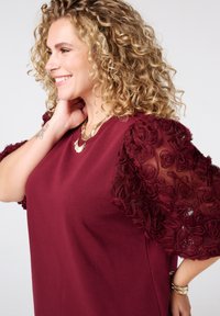 Haut bordeaux avec des manches bouffantes en tissu floral tissé, col rond et colliers superposés. Tissu doux avec des détails complexes aux épaules.