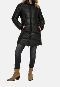 Zwarte gewatteerde jas met een hoge kraag, ritssluiting en zijzakken. Gedraaid over donkere spijkerbroeken en tan suède enkellaarsjes. Gladde, glanzende textuur.