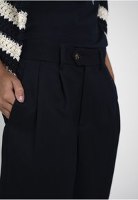 Main dans la poche portant un pantalon sombre taille haute avec des plis et un bouton marron, associé à un top tricoté rayé bleu marine et blanc.