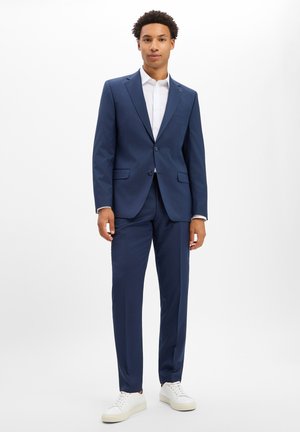 Man standing wearing a navy blue suit with a white shirt and white sneakers against a plain white background.