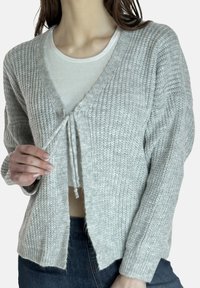 Grijze gebreide cardigan met een diepe V-hals, striksluiting, lange mouwen en geribbelde textuur. Gecombineerd met een wit binnenlaagje en donkere spijkerbroek.