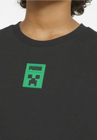 Maglietta nera a collo tondo con una toppa verde che presenta il logo Puma e un design di volto pixelato centrato sul petto.