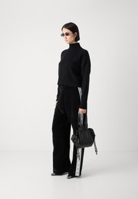 Zwarte korte trui met hoge hals; gecombineerd met zwarte fluwelen wijde broek met een witte zijstreep. Model houdt een zwarte handtas vast.