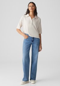 Kurzärmeliges, hellbeiges Poloshirt mit Knopfleiste, kombiniert mit hochgeschnittenen, weit geschnittenen blauen Jeans und weißen Schuhen.