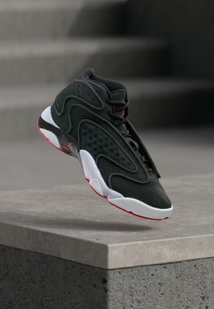 Zwart-rode hoge sneaker met witte zool die boven een betonnen rand zweeft met wazige trappen op de achtergrond.