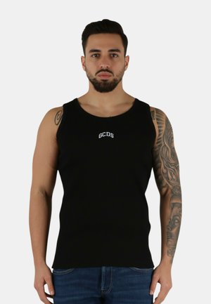 Uomo con barba e tatuaggi indossa una canotta nera GCDS e jeans blu, in piedi contro uno sfondo bianco uniforme.