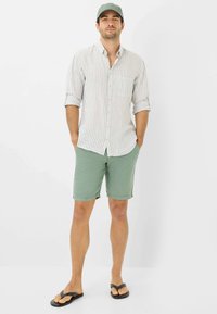 Outfit informale composto da una camicia a righe beige e bianca, pantaloni corti verdi e ciabatte nere, completato da un cappellino verde abbinato.