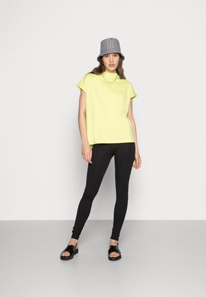 T-shirt amarelo de manga curta, com decote redondo, combinado com leggings pretas. Modelo usa sandálias pretas e um chapéu de balde xadrez preto e branco.