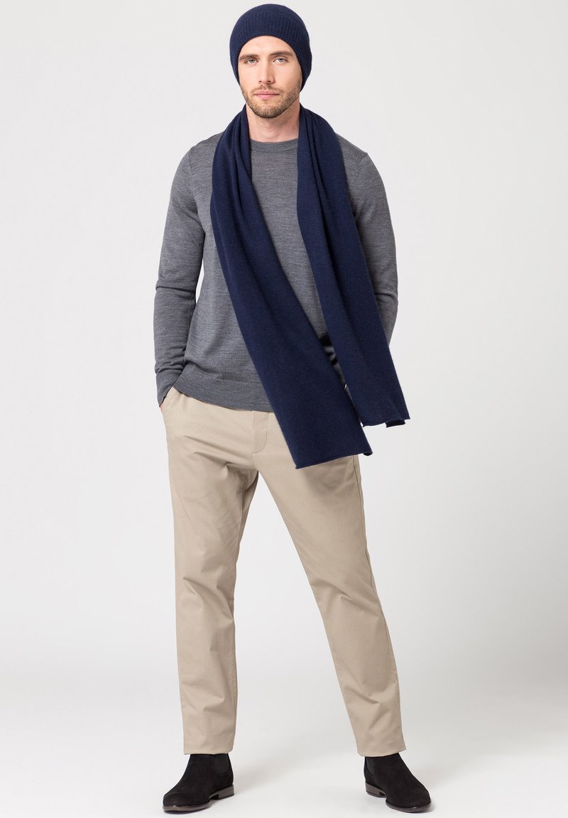 Bonnet en tricot marine et écharpe assortie, chemise à manches longues grise, pantalon beige et bottines noires. Texture douce, design décontracté.