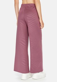 Wijdbeens pantalons in een gestructureerde stof in stofroze hebben een golvend patroon en een elastische tailleband, gecombineerd met witte sneakers.