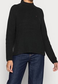 Zwarte geribbelde gebreide trui met een hoge hals, voorzien van gestructureerde zijaccenten en lange mouwen, gecombineerd met blauwe jeans met wijde pijpen.