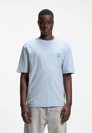 Jonge man met een lichtblauw textuur kortärmelig overhemd met een klein blauw "H²O"-logo op de borst en lichtgrijze broek.