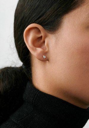Detail ucha osoby nosiacej malý strieborný náušnicu v tvare kvapky, s tmavými vlasmi stiahnutými dozadu a viditeľným čiernym rolákom.