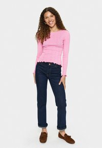 Vrouw met krullend haar, in een roze lange mouw top, donkere blauwe opgerolde jeans en bruine mokassins, die glimlacht tegen een witte achtergrond.