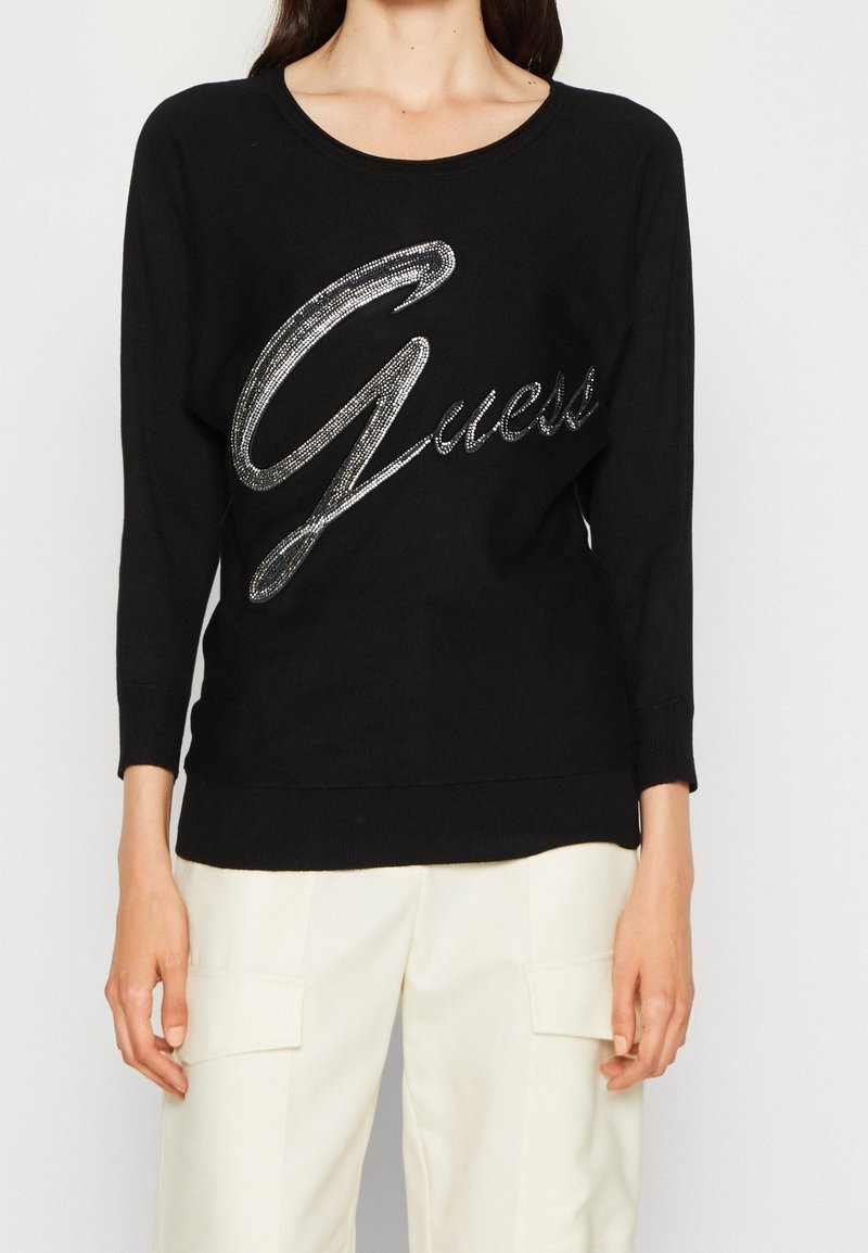 Femme portant un pull noir à manches longues avec un grand logo argenté "Guess" et un pantalon blanc avec poches avant, debout sur un fond blanc.