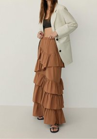 Jupe maxi évasée marron en tissu léger, assortie à un crop top gris et un blazer crème. Des sandales noires à lani�ères complètent le look.
