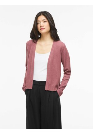 Kvinne med langt mørkt hår iført en rosafarget cardigan over en hvit topp og svarte vide bukser, står med hendene i lommene.