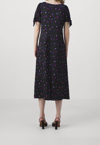 Kvinna klädd i en svart midiklänning med lila och rosa blommönster, korta knutna ärmar och svarta klacksandaler, stående vänd bort.