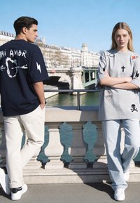 Deux jeunes adultes portant des T-shirts graphiques oversize et des pantalons clairs se tiennent près d'une balustrade en pierre surplombant une rivière et un pont.