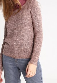 Rosa gestrickter Pullover mit Rundhalsausschnitt und langen Ärmeln, mit einer strukturierten Oberfläche und dezenten metallischen Fäden. Kombiniert mit hellblauen Jeans.