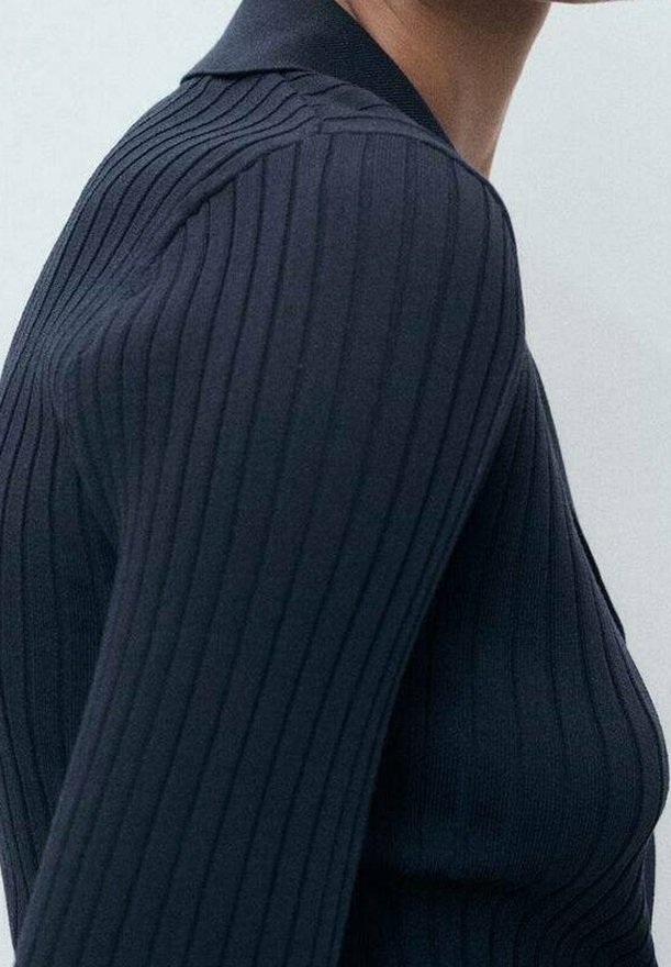Personne portant un pull côtelé bleu marine ajusté avec un col montant, vue de profil sur un fond clair uni.