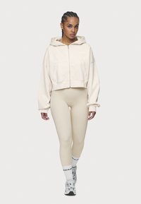 Cropped beige hoodie with a front zipper and pocket, paired with beige leggings, white socks, and gray sneakers. Smooth texture, casual design.